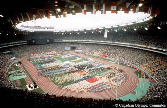 A colour photograph depicts the inside of Olympic Stadium, Montréal. The athletes of the competing nations assemble on the infield of the stadium. The flags of the competing nations hang from the rafters.