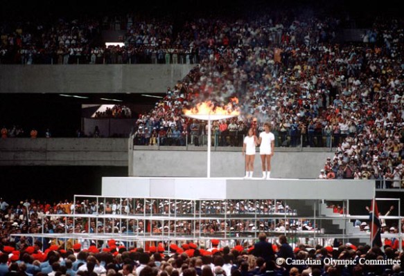 A colour photograph depicts two young people standing on a raised platform in a crowded stadium. They each have a hand on a torch which they are holding aloft. Beside them is a large ceremonial cauldron that is lit with a flame.