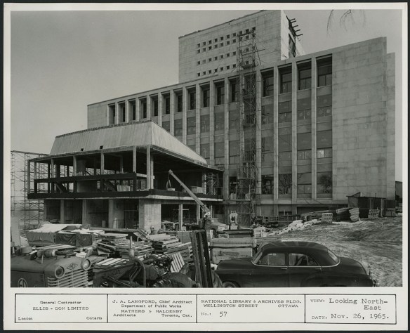 A black-and-white photograph showing a construction site with a partially finished building. 