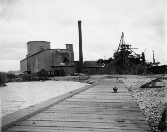 A black-and-white photograph showing a long wooden dock by the shore and grain elevators in the background