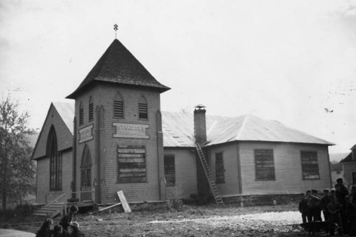 A black-and-white photograph showing a boarded-up church with construction material scattered around it