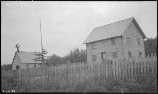 A black-and-white photograph showing two small abandoned buildings, one possibly a school, behind a white picket fence