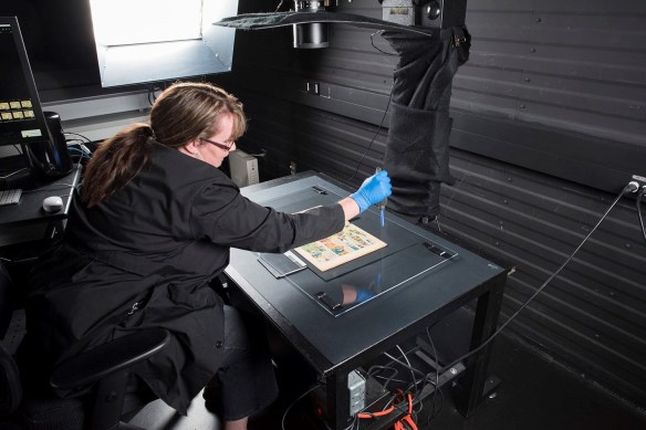 A comic book is placed on a flat black surface underneath a sheet of Plexiglas. A woman leans over the surface, using an anti-static blower to remove dust from the Plexiglas. The lens of a camera is visible above the table.