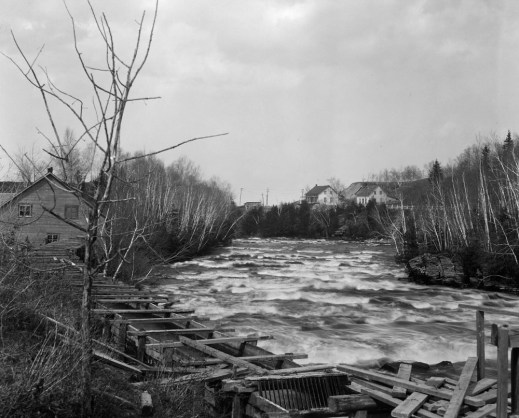 A black-and-white photograph showing a river with a heavy current, trees on both sides, and an old mill to the left in the background