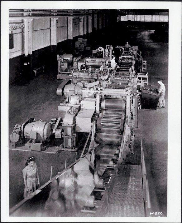 Black and white photograph showing workers operating a machine used to roll the steel and make it into panels. 
