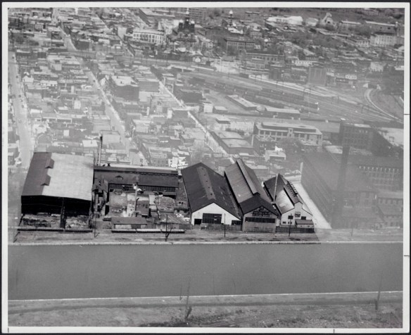 Black and white photograph showing a mill beside a canal. Other factories and railway tracks for transporting steel materials can be seen in the background.