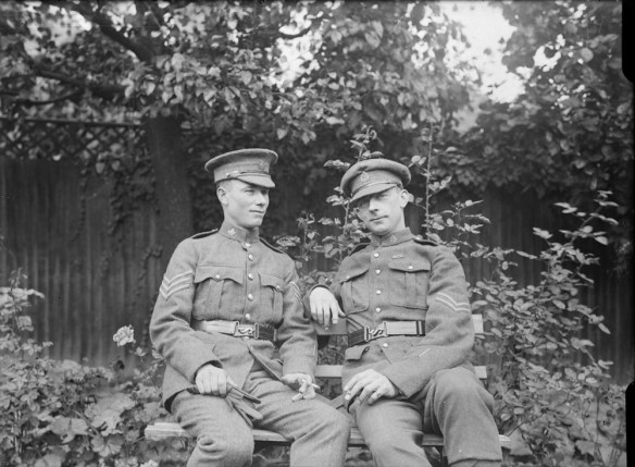 A black-and-white photograph of two soldiers in uniform sitting on a bench. The man on the right is looking directly at the camera with a slight smile.