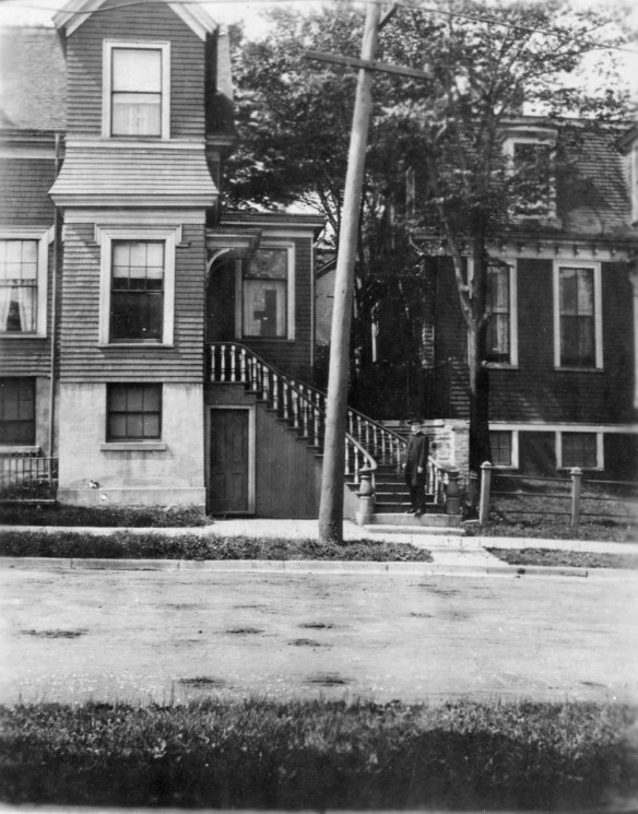 Black-and-white photograph featuring Dr. Edward M. Saunders, standing on the front staircase of a three-story Victorian style house. Dr. Saunders is wearing a black clerical suit and a black hat. The photograph was taken from across a residential street. 