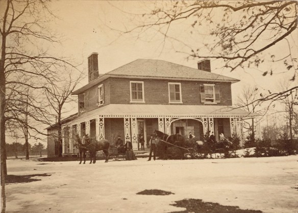 A black and white image of a house with melting snow all around. In front of the house are two horse-drawn sleighs with people around them.