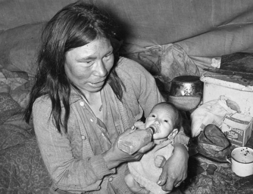 Black and white photograph of a gaunt looking Inuit woman and child sitting in a small tent with cooking supplies in the background. The woman is feeding the baby with a rectangular bottle.