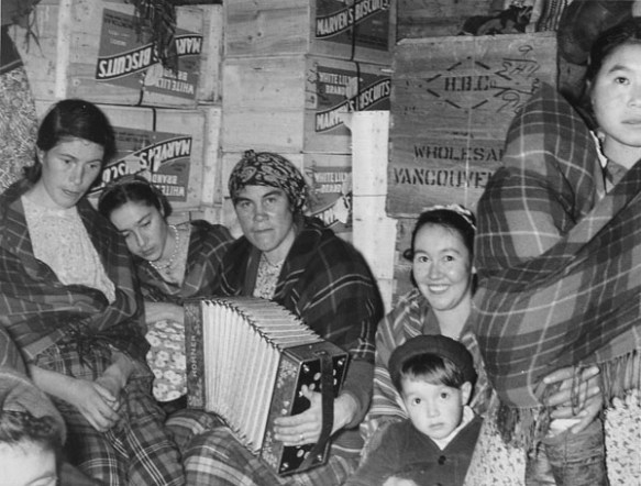 Black and white photograph of five women, four of them seated and one on the far right standing with a baby wrapped in a plaid shawl. The woman in the centre of the photograph is playing an accordion, and the woman to her left has a young boy in her lap. Behind them are several wooden crates labeled “Marven’s Biscuits” and one marked “H.B.C. Wholesale Vancouver.”
