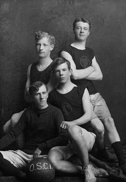 A black-and-white photograph showing four young men posing around a basketball.