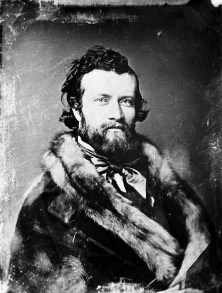 A black-and-white photograph of a young, bearded man looking directly at the camera. He is wearing a fur coat and a very dapper cravat.