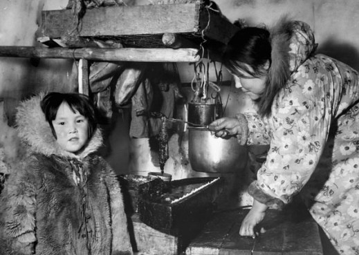 Black-and-white photograph of an Inuit woman inside an igloo wearing a floral print parka and tending a seal oil lamp, with a young Inuit child wearing a fur parka. 