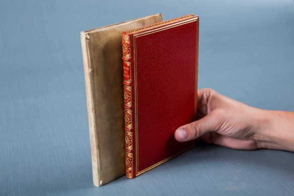 A hand holds two books upright against a grey-blue background. The book on the left is in a very plain beige cover. The book on the right is in a red cover decorated with gold.
