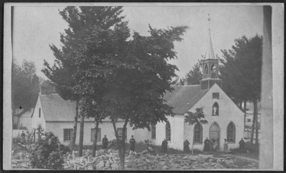 Black-and-white photo of a church set among trees with church goers standing along the walls.