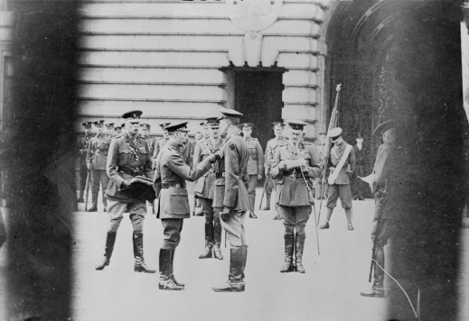 A black and white photograph of a man pining an award on another man’s pocket. Another man is reading the citation while a third man is carrying a case. In the background, rows of soldiers are standing at ease.