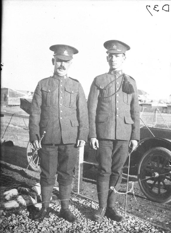 A black-and-white photograph of two soldiers standing side by side in front of an automobile.