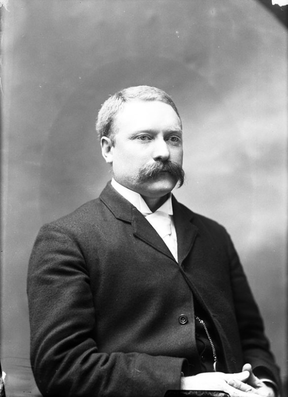 Black-and-white photo of a man with a moustache, wearing a suit and a white shirt.