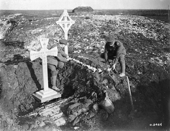 A black-and-white photograph of two men adorning a makeshift grave with white stones in a desolate landscape that has patches of snow and frost on the ground. The grave is marked by a cross with the words “L.S. [Lance-Sergeant] E.W. Sifton, VC” and adorned with a maple leaf. Beside the grave is a larger cross with the words “RIP Canadian soldiers killed in action 9-4-17.”