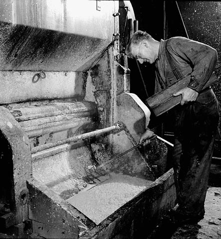 A black-and-white photograph of a man leaning over a grinder machine holding a plank of wood in his hands. 