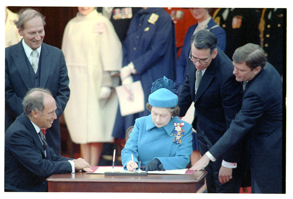 Woman in blue sitting at a desk signing a paper. Four men in suits surround her; two leaning over the desk, one sitting to the side, and the fourth standing back to the side.