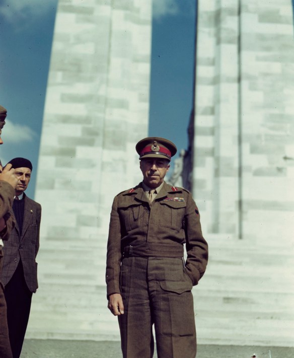 A colour photograph of man standing in front of a large stone structure. Two people are on the left side of the photograph, one is in uniform and mostly cut off and the other is wearing a vest, sweater and beret.