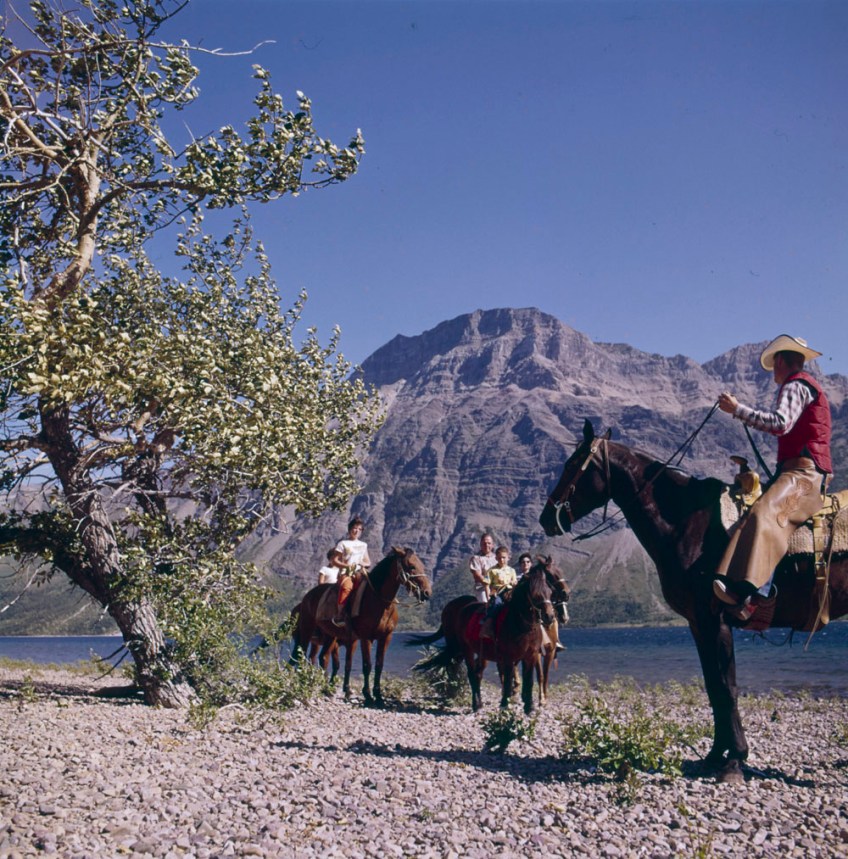 A colour photograph of a group of people on horseback by a river with a mountain peak in the background. One is dressed in “cowboy” attire and appears to be leading a family on a trail.