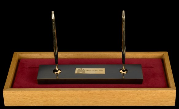 Two gold and black pens standing upright in a gold and black pen stand resting on a velvet pad in a wooden box.