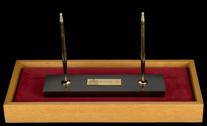 Two gold and black pens standing upright in a gold and black pen stand resting on a velvet pad in a wooden box.