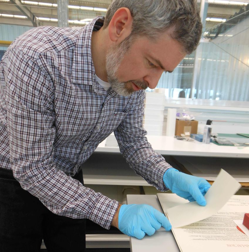 A colour photograph of a man wearing blue gloves bent over a table and arranging papers.
