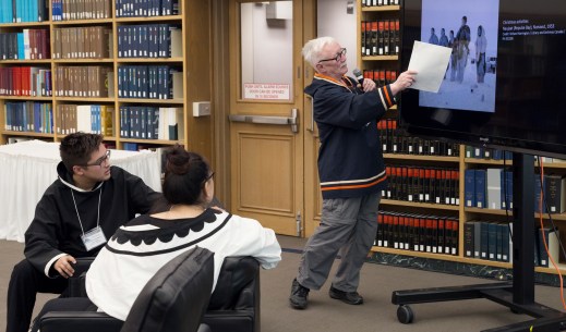 A colour photograph of an older Inuit man wearing a traditional Inuit jacket who is standing in front of a monitor pointing to an image on the screen. Two young Inuit are watching and listening closely.