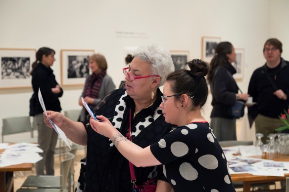 A colour photograph of two Inuit women looking closely at a picture. The older one holds the picture while the younger one takes a photo on her iPhone of it. Behind them are people talking and tables covered with papers.
