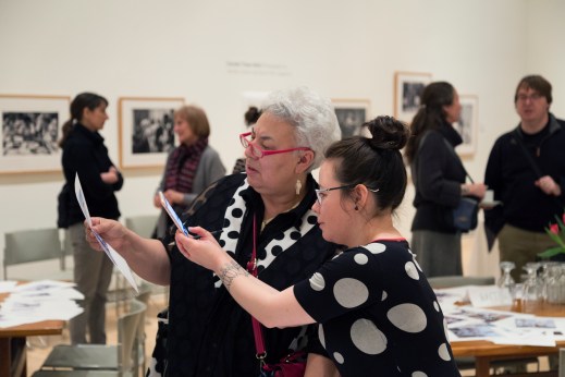A colour photograph of two Inuit women looking closely at a picture. The older one holds the picture while the younger one takes a photo on her iPhone of it. Behind them are people talking and tables covered with papers.
