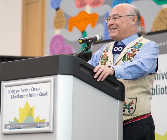 A colour photograph of a man from the Métis Nation wearing a beaded vest standing at a podium. 