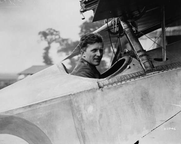 A black-and-white photograph of a man sitting in the open cockpit of an airplane looking at the viewer. 