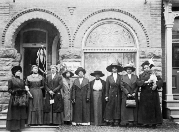 Black and white photograph of nine women wearing dresses, coats and hats standing in front of a residential building. 