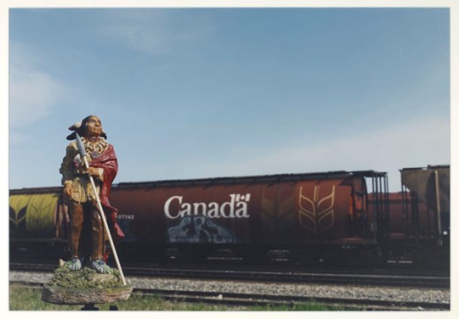 First Nations figure set in front of a train marked “Canada” with grain graffiti on its side.
