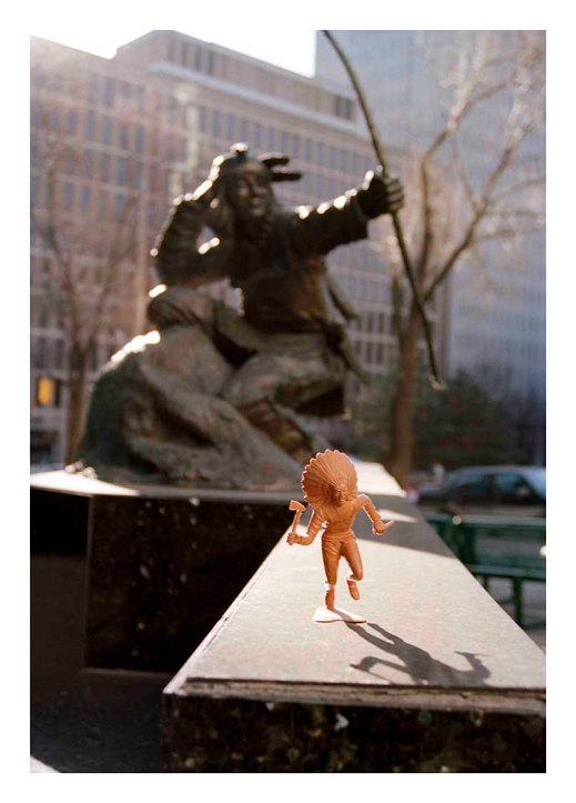 Small light-brown First Nations figure set in front of Indian hunter statue. Tall office buildings and trees can be seen in the background.