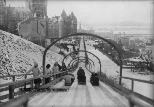 Black and white photo of tobogganists sliding past a group of people walking up to the top of the run.