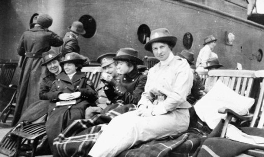 A black-and-white photograph of a group of four women sitting on deck chairs with blankets. Three of the sisters are wearing the dark overcoat while one is wearing a lighter coloured jacket. A soldier can be seen slightly in the middle of the group.