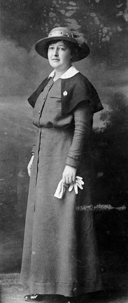 A black and white photograph of a woman in a nursing sister uniform with the cape, pin, hat and white gloves. 