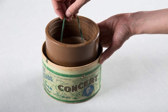 A colour photograph of a wax cylinder being pulled out of its protective cardboard container by a string that is attached on the inside of the cylinder. On the cardboard, the word “Concert” appears in uppercase letters, while above, in smaller print, can be read “National Phonograph Co, New York, U.S.A.” “Made at the Edison Laboratory, Orange, N.J.” is visible below.
