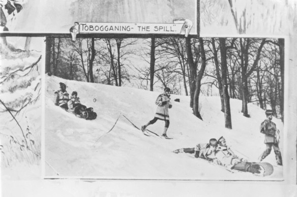 A black-and-white photograph of a winter scene of people on toboggans and others on snowshoes descending a hill. 