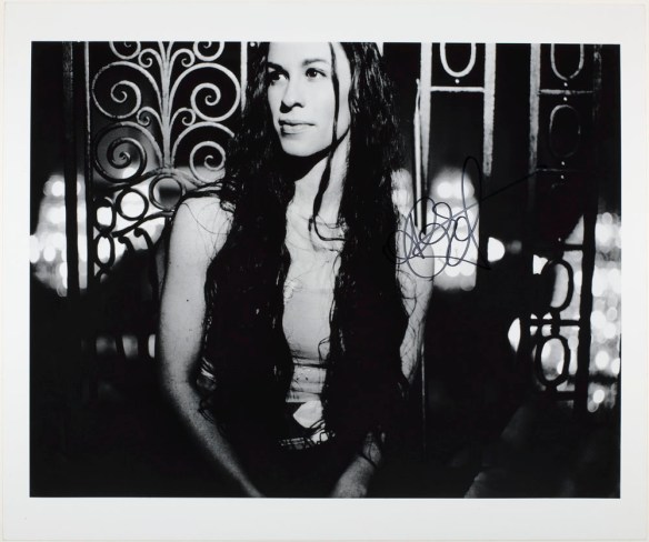 A black-and-white photo of a woman with long hair leaning against a wrought iron fence.