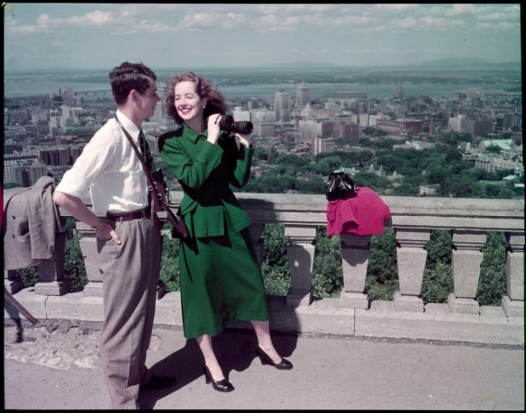 A colour photograph of a couple standing with binoculars looking over the city on the edge of a lookout. 