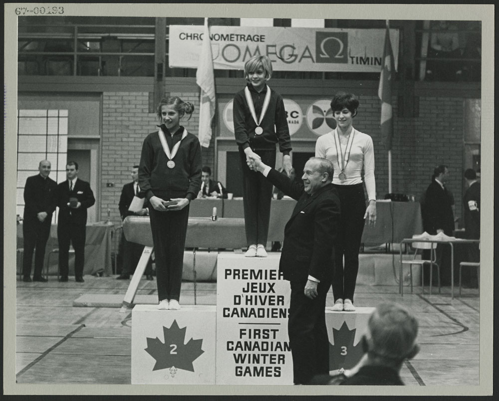 A black-and-white photograph of a podium on which three young women wearing medals are standing. A man is shaking hands with the gold medallist.