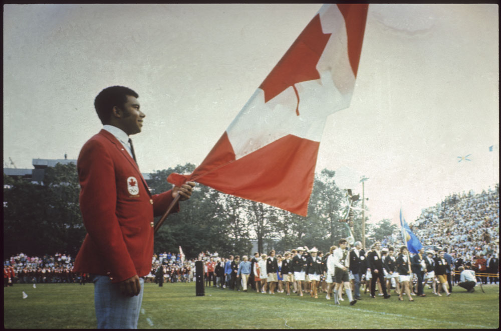 Colour photograph of a man in a red jacket carrying the Canadian flag while athletes enter the stadium.