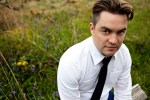 A colour photograph of a young man wearing a white shirt and tie, sitting in a field.