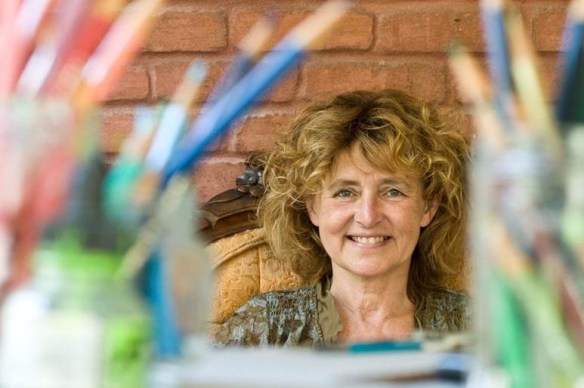 A colour photo of a seated, smiling woman. Blurred pencil crayons can be seen in the foreground.